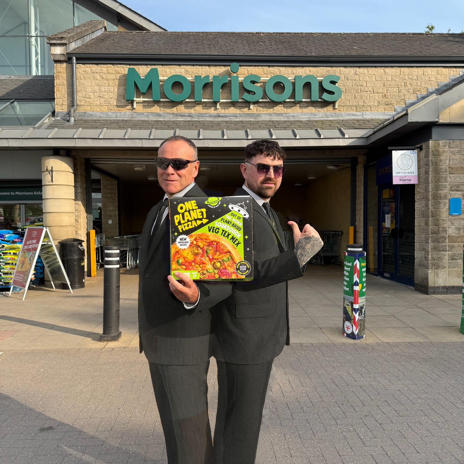 Two men in suits holding a one planet pizza box in front of a Morrisons store.